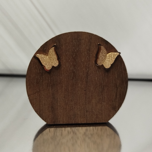 Wooden earring display with gold butterfly earrings on a reflective surface.